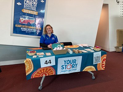 A smiling woman with short, curly blonde hair sits behind a table for Your Story Disability Legal Support